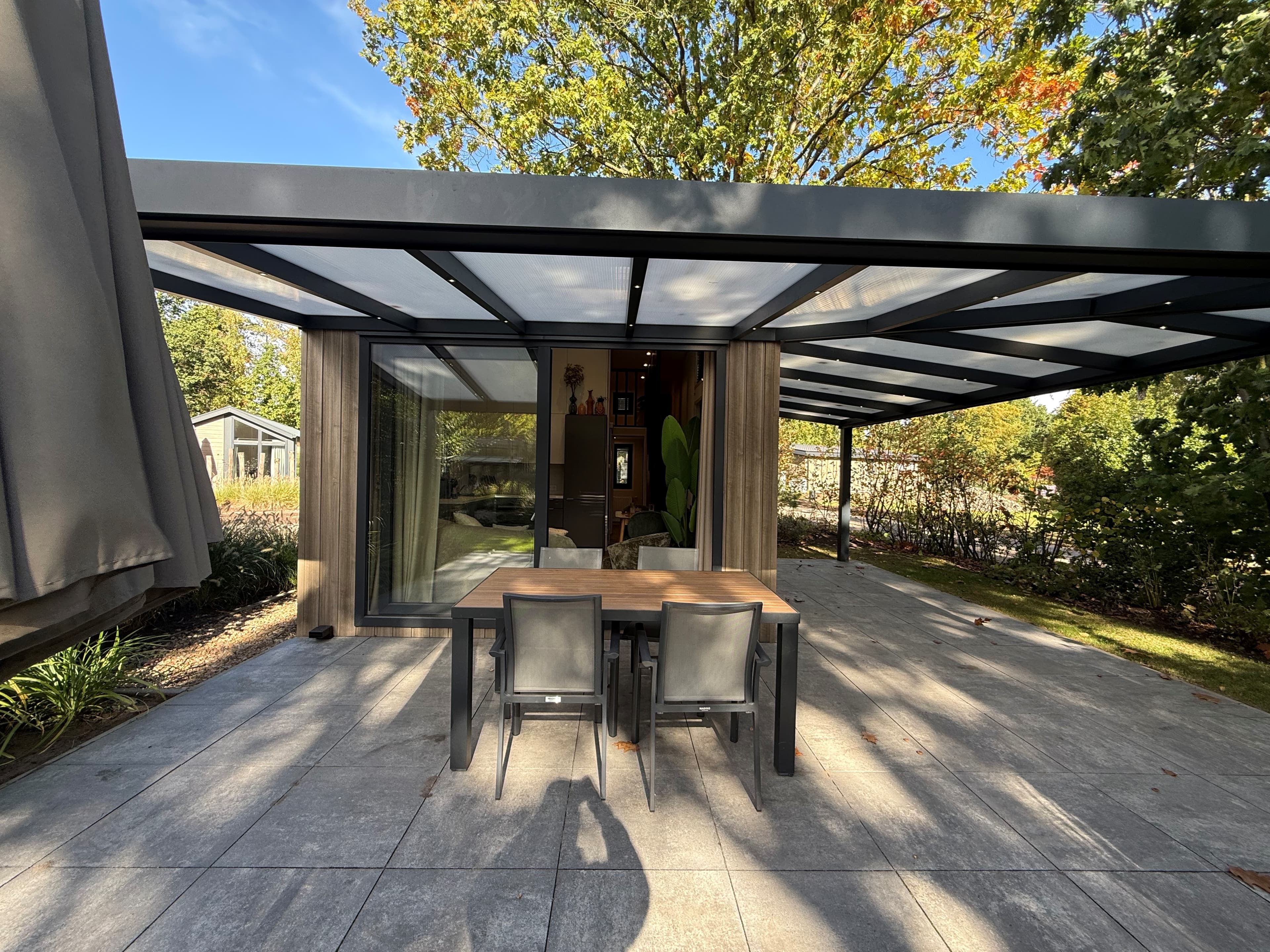 Deeltuin met een houten tafel en stoelen onder een overkapping bij een recreatiewoning, omringd door groen.