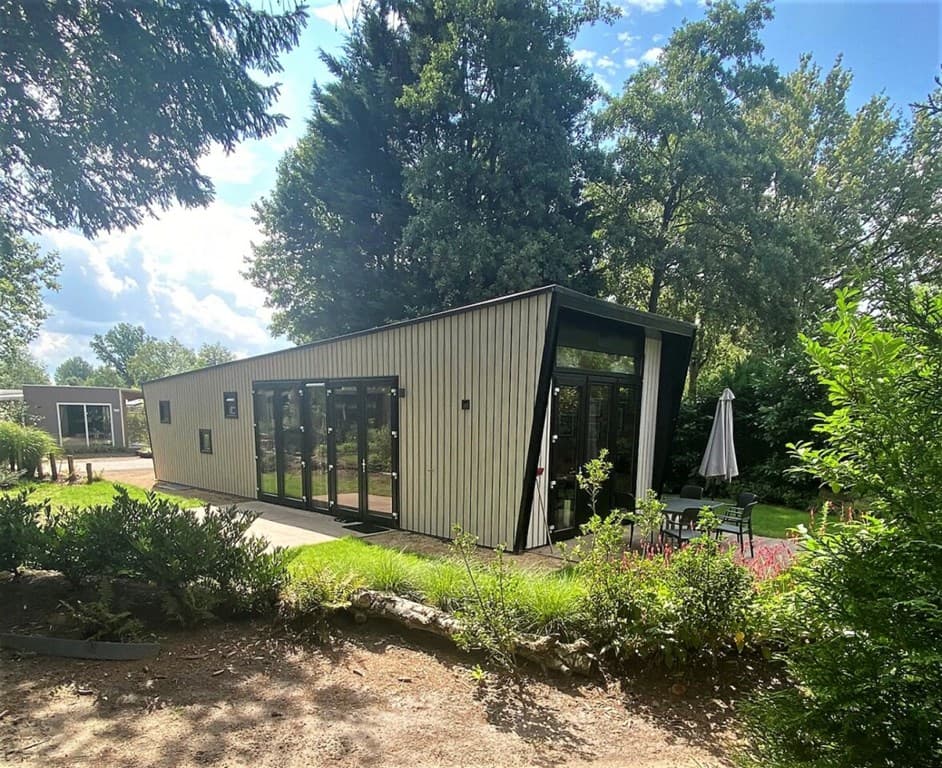 Moderne vakantiewoning met grote ramen, omgeven door groene bomen en een terras met parasol en tuinmeubilair.