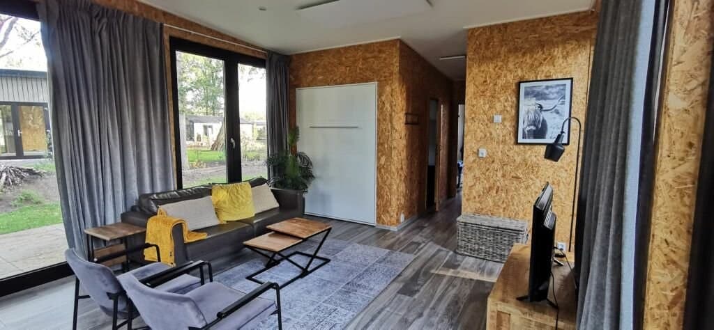Woonkamer met houten wand, bank, fauteuils, salontafel en tv, grote ramen met uitzicht op de tuin.