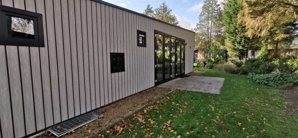 Moderne woning met houten gevel en grote glazen schuifpui, omgeven door groene tuinen en bomen
