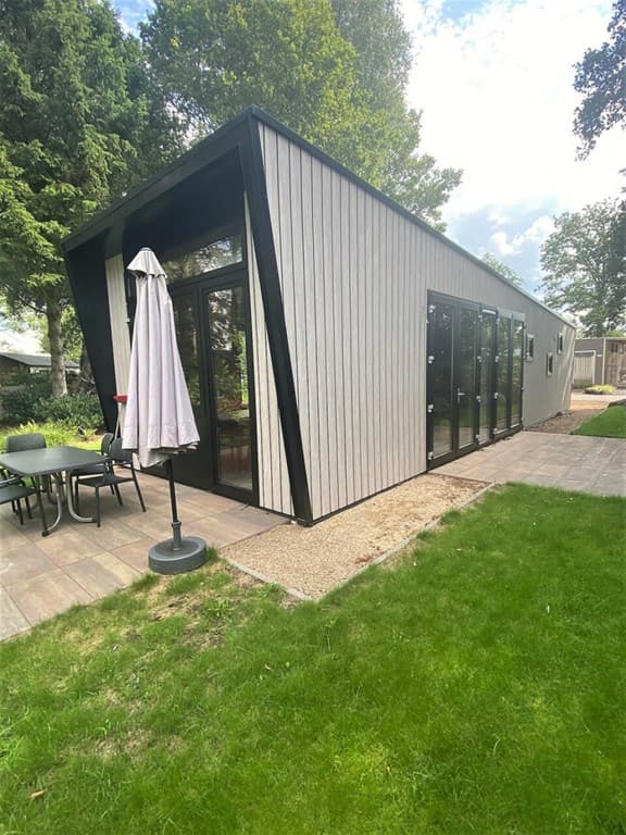 Moderne vakantiewoning met schuin dak en grote ramen, omgeven door gras en een terras met parasol en tuinmeubilair