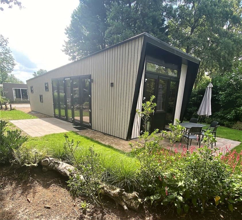 Moderne vakantiewoning met grote ramen, terras met stoelen en parasol, omringd door groen landschap.