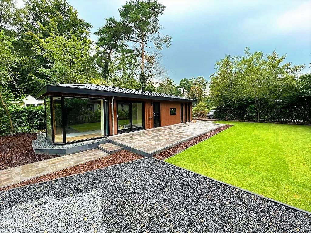 Moderne vakantiewoning met grote ramen, omgeven door groen gras en bomen, met een betegeld terras en grindpad.