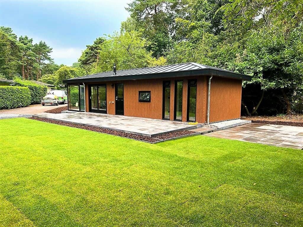 Moderne vakantiewoning met houten gevel, grote ramen en betegeld terras, omgeven door groen gras en bomen