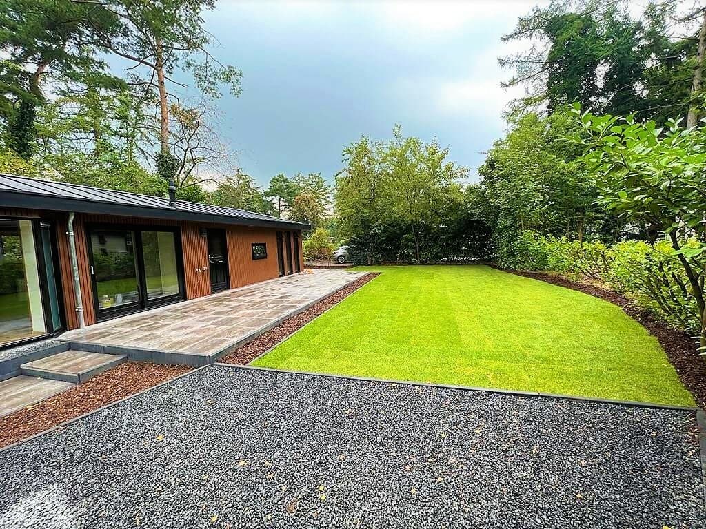 Moderne woning met groene tuin, betegeld terras en grindpad omringd door bomen en struiken