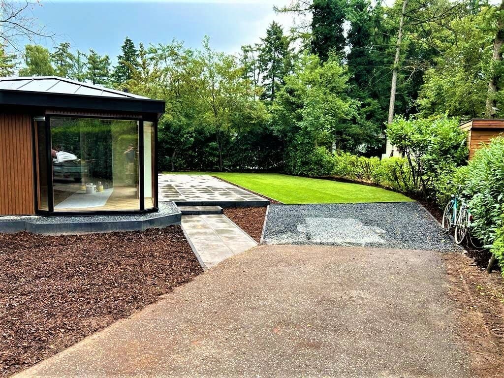 Tuin met een betegeld pad, gazon, en een moderne woning met grote schuiframen aan de zijkant.