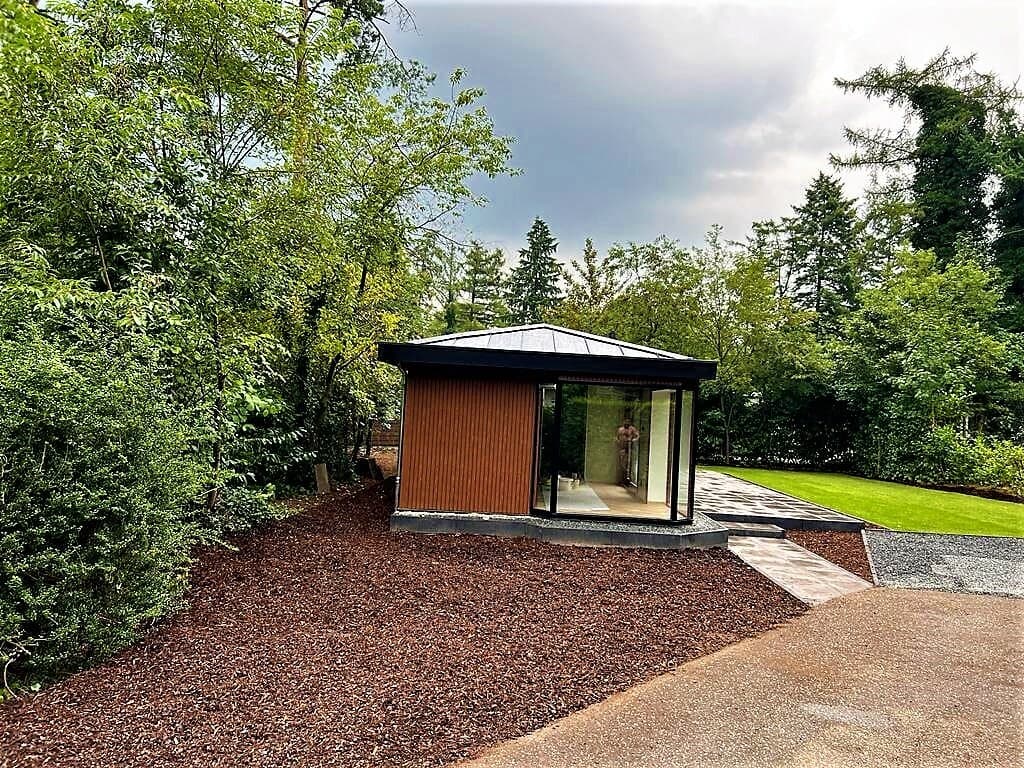 Moderne vakantiewoning met grote raampartijen, omgeven door bomen en een gazon met grindpad.