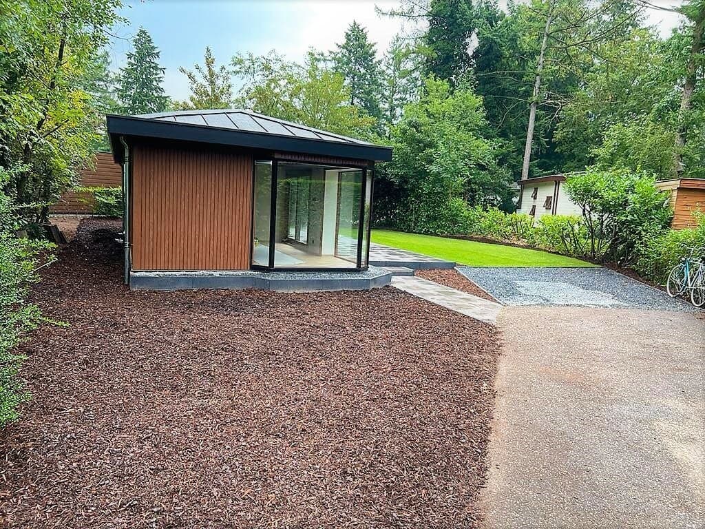 Moderne woning met grote glazen pui, omringd door bomen en een betegelde oprit met grind en groen gras.