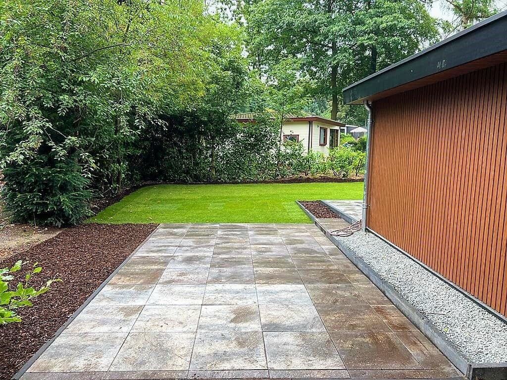 Tegeltuin met groen gazon en omliggende bomen, naast een moderne woning met houten gevel aan EuroParcs De Achterhoek.