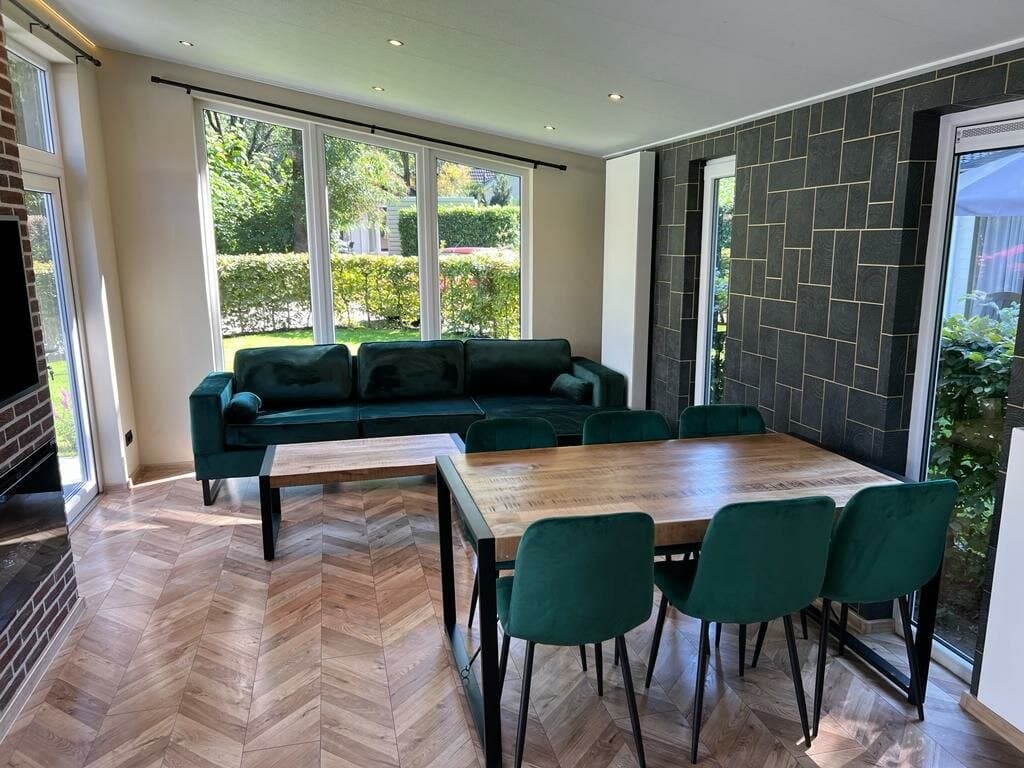 Huiskamer met groene bank en houten eettafel met zes stoelen, grote ramen met uitzicht op de tuin.