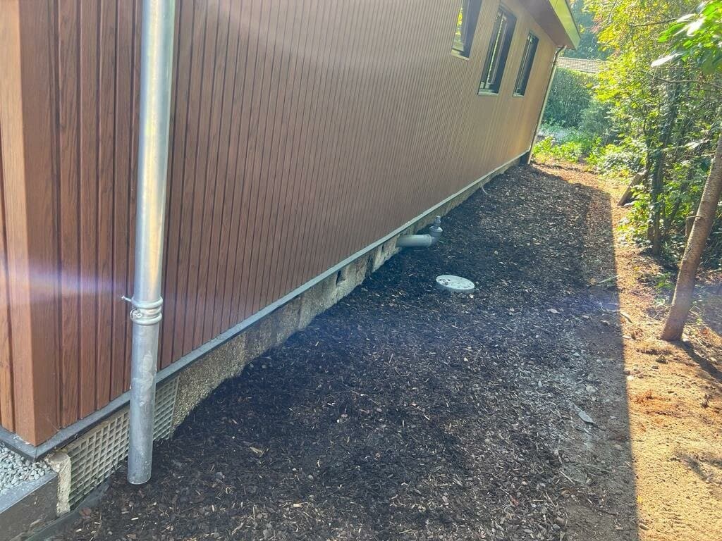 Zijaanzicht van een woning met houten gevelbekleding en een aangelegde tuin met houtsnippers langs de fundering.