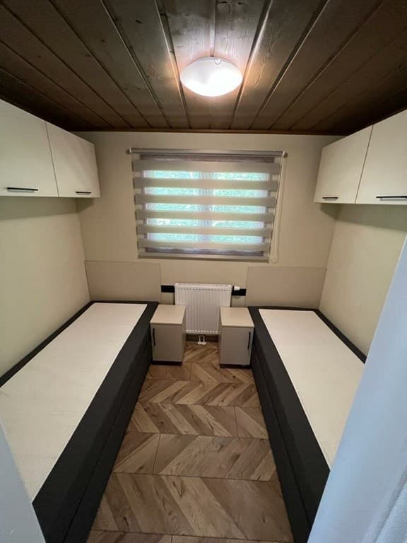 Slaapkamer met twee aparte bedden, nachtkastjes en een raam met jaloezieën, houten vloer en plafondlamp.