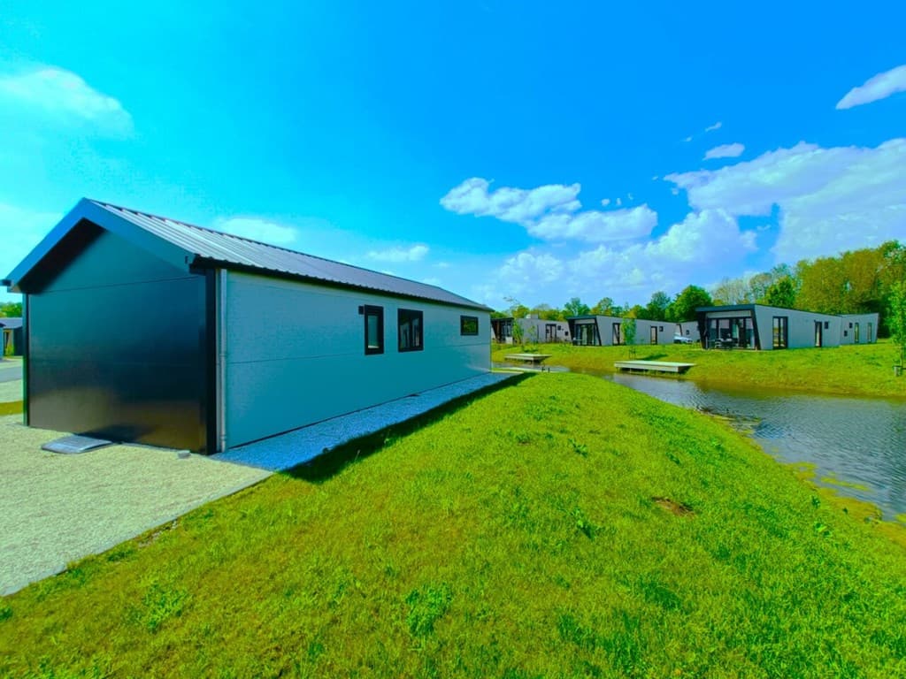 Moderne vakantiewoning aan een waterrand met groene ruimte en andere woningen op de achtergrond onder een blauwe lucht