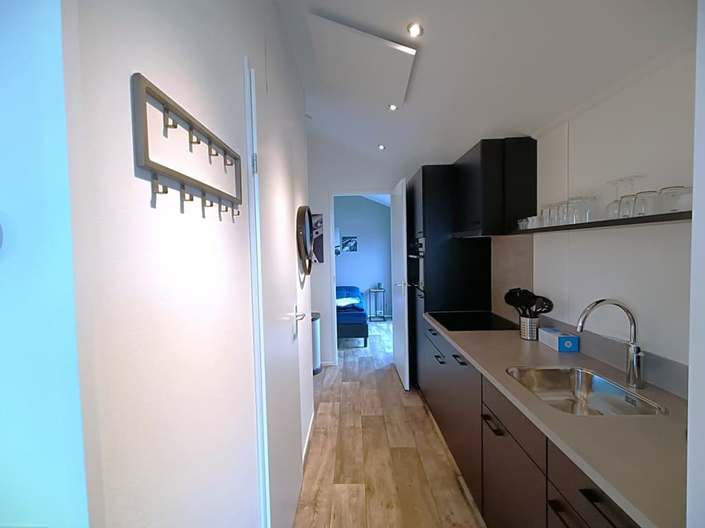 Smalle gang met keuken aan de rechterzijde en een deur die leidt naar een woonkamer aan het einde van de gang.