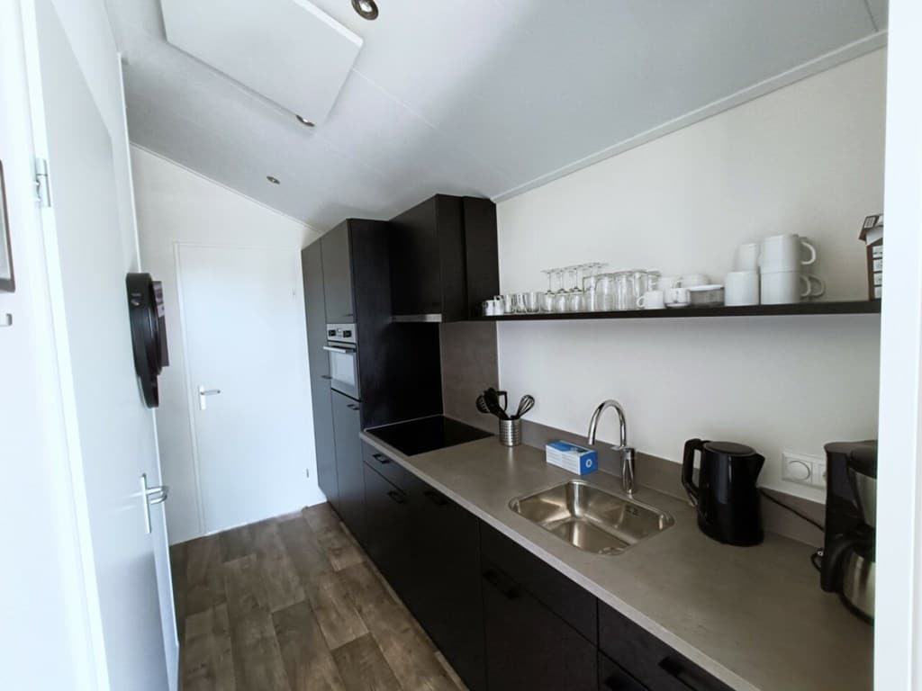 Keuken met zwarte kasten, aanrecht met spoelbak en wandplanken met glazen en servies.