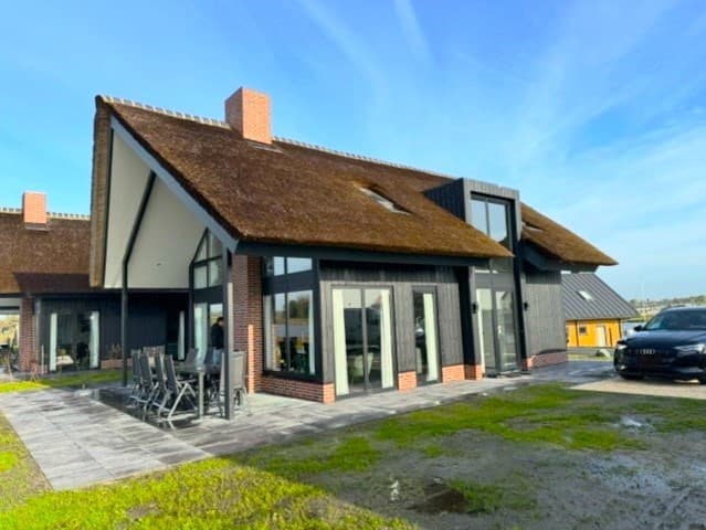 Twee-onder-een-kap woning met rieten dak, grote ramen en terras met tuintafel en stoelen, gelegen op een groene kavel.