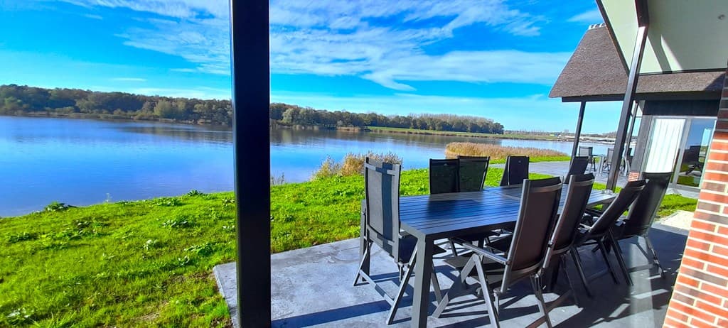 Terras met grote eettafel en stoelen, uitzicht op het meer en groene omgeving met bomen aan de oever