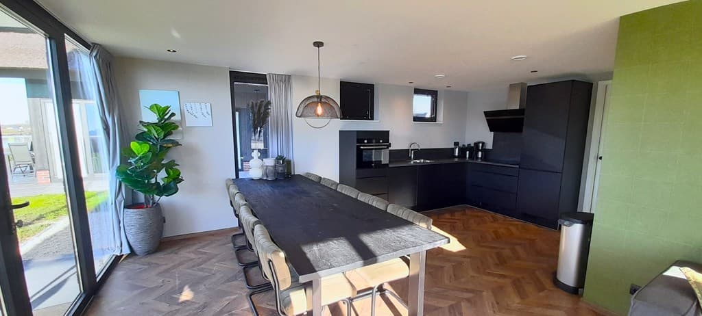 Moderne keuken en eetruimte met lange tafel, zwarte kasten en grote ramen met uitzicht op de buitenruimte.
