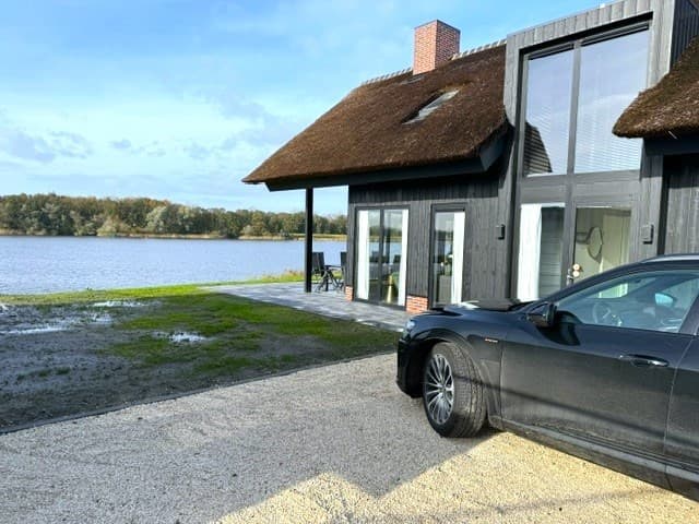 Zwarte woning met rieten dak aan het water, uitzicht op een grasveld en een grijze auto voor de deur.