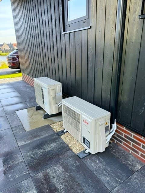 Buitenaanzicht van een woning met twee airconditioningunits op een betegeld terras en een gevel van zwarte houten planken