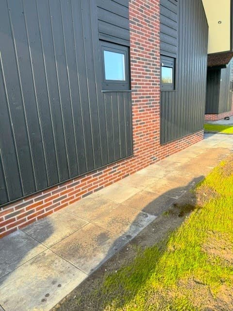 Zijaanzicht van een woning met houten gevel en metselwerk, voorzien van kleine ramen en tegels op de grond