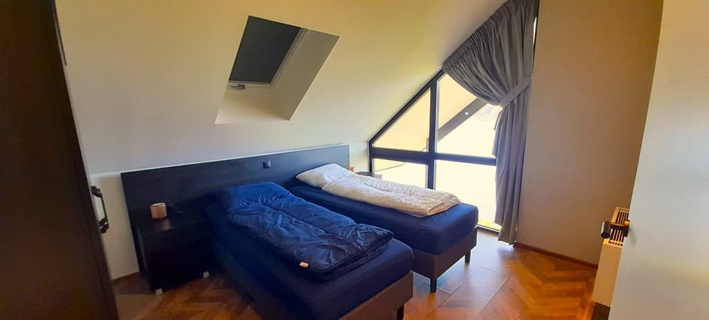 Slaapkamer met twee losse bedden, grote ramen en een schuin plafond met een gordijn.