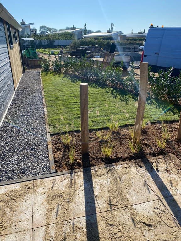 Tuin met een gazon, grindpad en houten palen naast een woning, groene omgeving en een witte bestelauto op de achtergrond