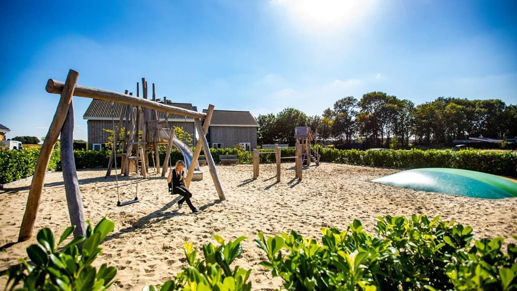 Speelplek met zand, schommels en klimtoestellen, omringd door struiken en huizen onder een blauwe lucht.