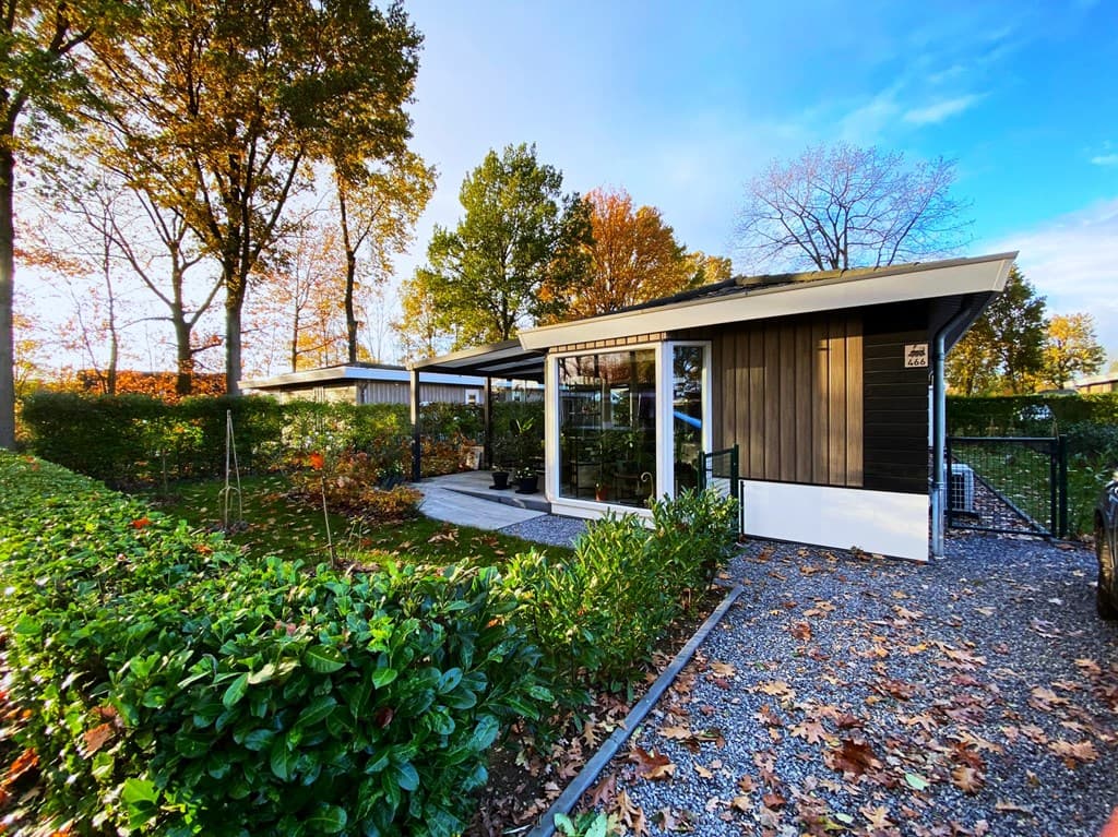 Moderne woning met een plantenborder en terras, omgeven door bomen met herfstbladeren en een zichtbare oprit.
