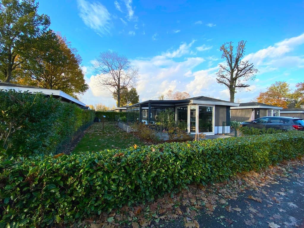 Vakantiewoning met groene tuin en haag, omgeven door bomen en andere woningen, onder een heldere lucht