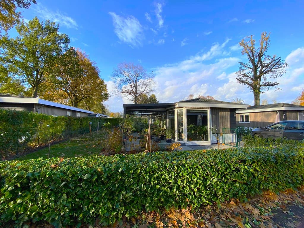 Buitenzicht van een moderne vakantiewoning omringd door groen en bomen, met een overdekt terras onder een blauwe lucht