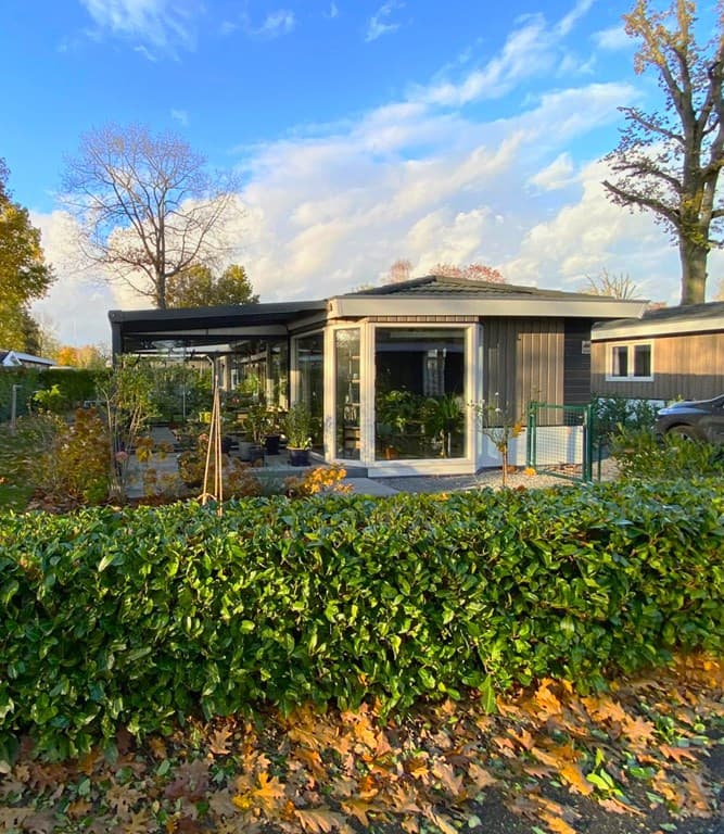Moderne vakantiewoning met grote ramen, omgeven door groen en een blauwe lucht met wolken.