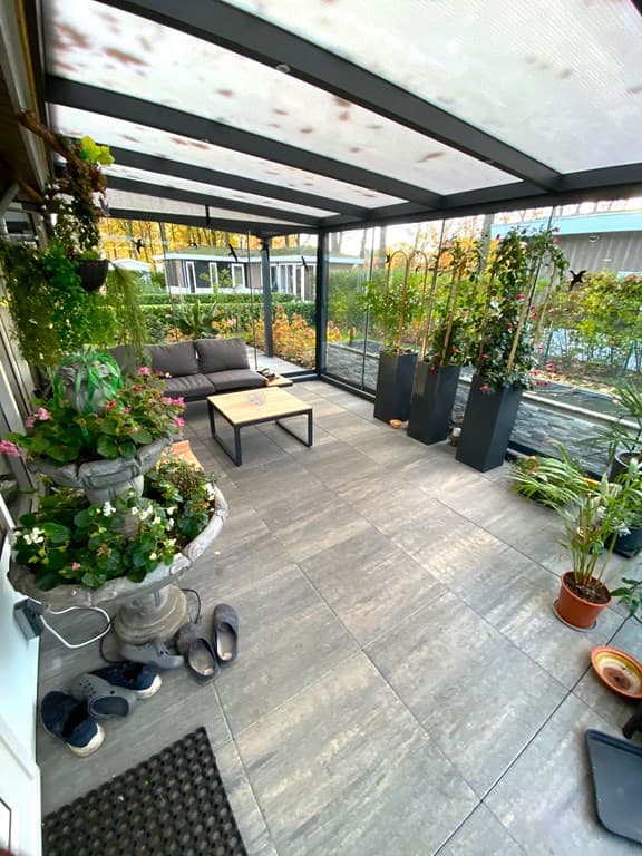 Veranda met een zithoek, planten, voorbijgang en een tegelvloer met potplanten en slippers op de grond.
