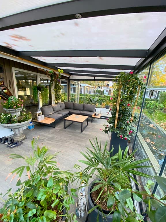Terras met moderne zithoek, planten in potten en glazen afscheiding, omringd door groen en rustige omgeving