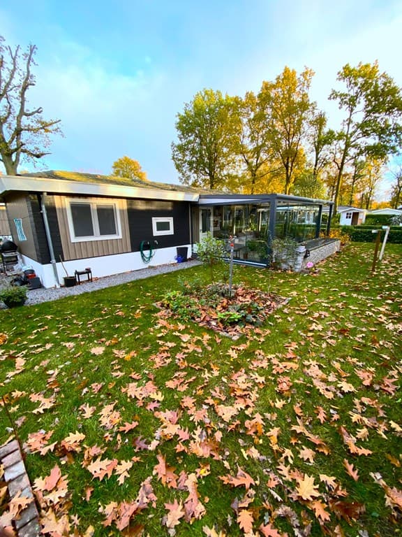 Vrijstaande woning met een overdekt terras, omringd door groene grasvelden en bladeren in herfstkleuren.