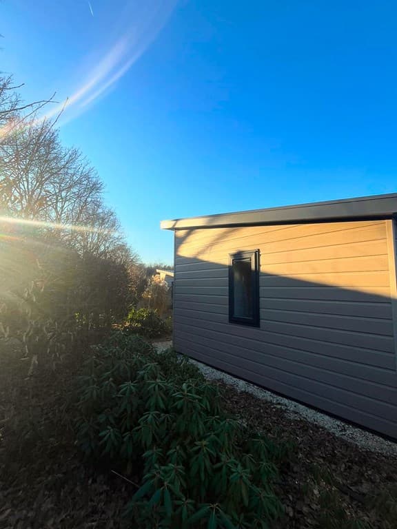 Zijaanzicht van een woning met een blauwe hemel en zonneschijn, omgeven door bomen en groene planten