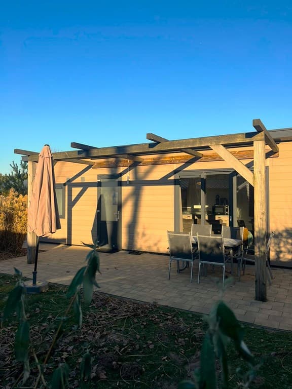Buitenzicht van een woning met een terras, houten overkapping en tuinmeubilair onder een blauwe lucht