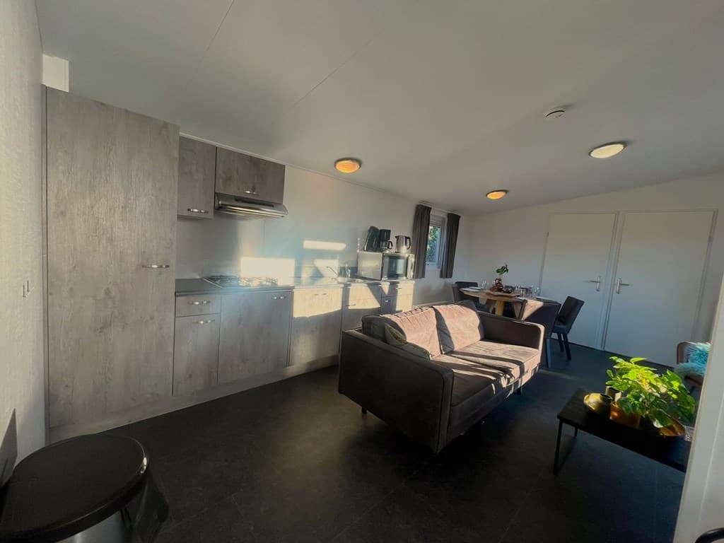 Lichte woonkamer met een grijze keuken, een bank en een eettafel met stoelen. Planten en een donkere vloer zijn zichtbaar.