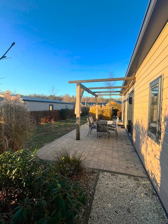 Terras met houten pergola en eettafel, omgeven door groen en struiken, met uitzicht op een open ruimte.
