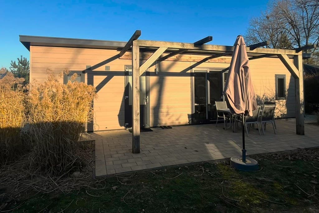 Moderne vakantiewoning met houten pergola, terras en tuinmeubelen, omgeven door gras en struiken.