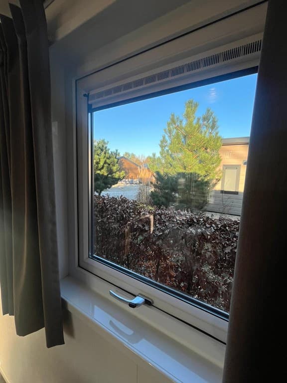 Uitzicht vanuit een raam met groene bomen en enkele woningen op de achtergrond, lichte gordijnen aanwezig.