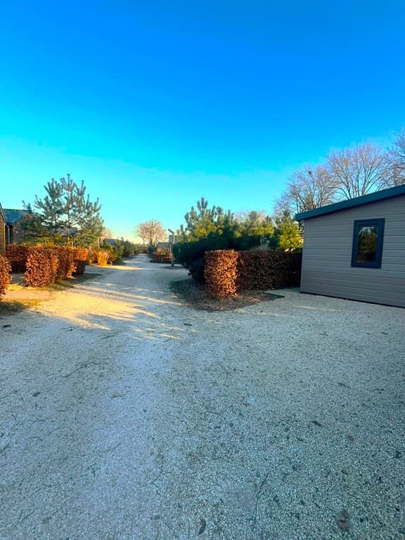 Zicht op de gravelweg omringd door groene struiken en bungalow aan de rechterzijde met helder blauwe lucht.
