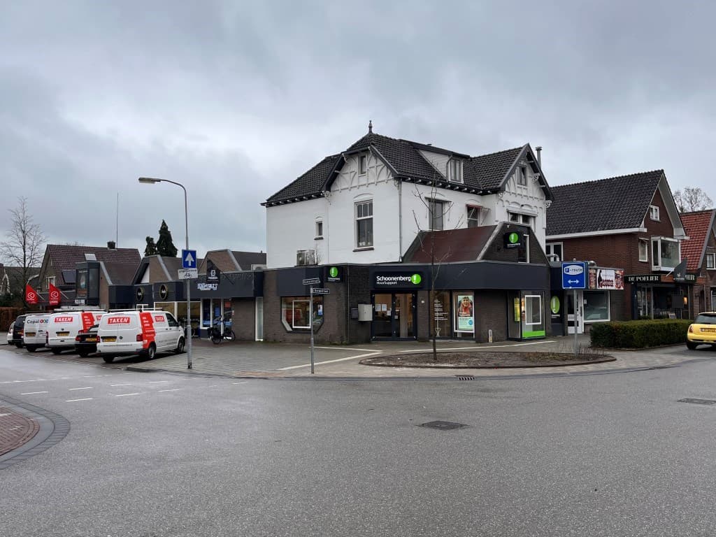 Hoekpand met verschillende winkels en een woning op de bovenverdieping, grijs weer en meerdere voertuigen zichtbaar op de str