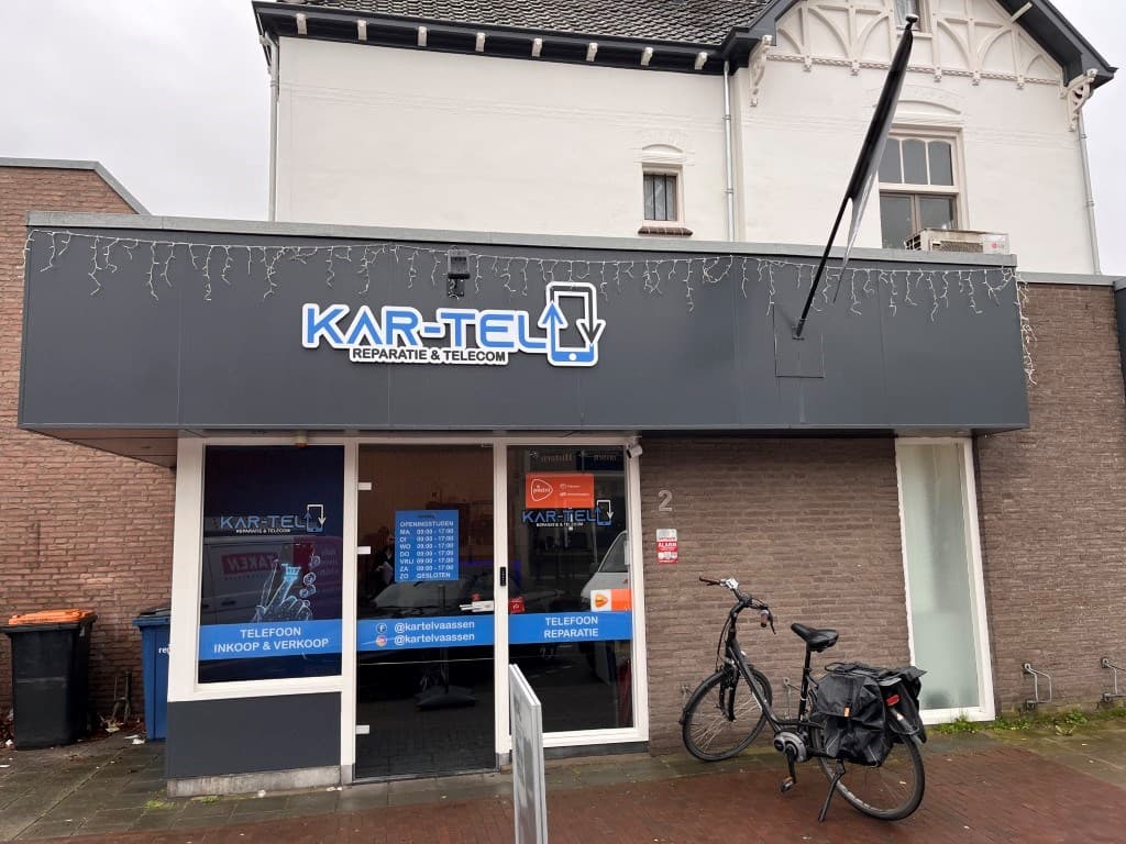 Winkelpand van Kar-Tel met een grijze gevel en blauwe logo, fiets staat naast de ingang, kerstverlichting aan de voorkant.