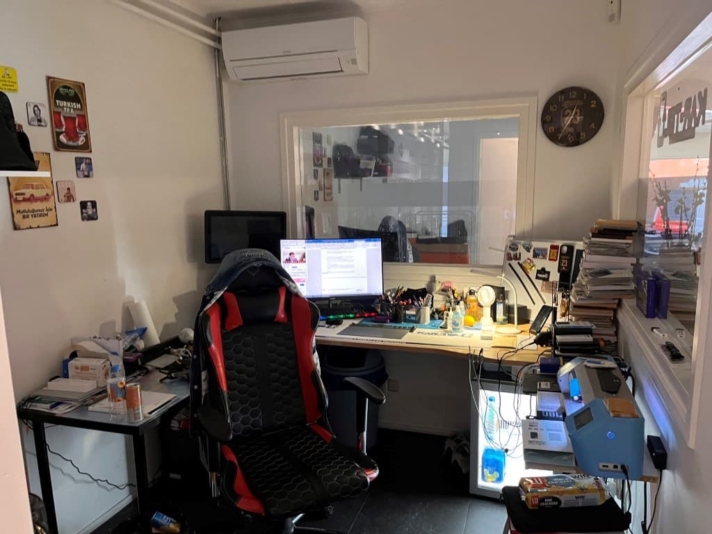 Kantoorruimte met een bureau, een computerscherm, een bureaustoel en verschillende stapels papieren en tijdschriften.