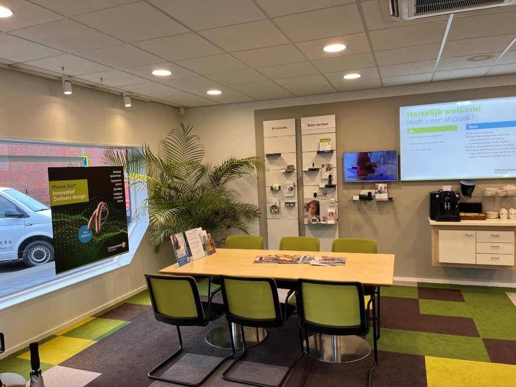 Informatieruimte met een grote tafel, groene stoelen en een plantenbak, voorzien van diverse brochures en een scherm.