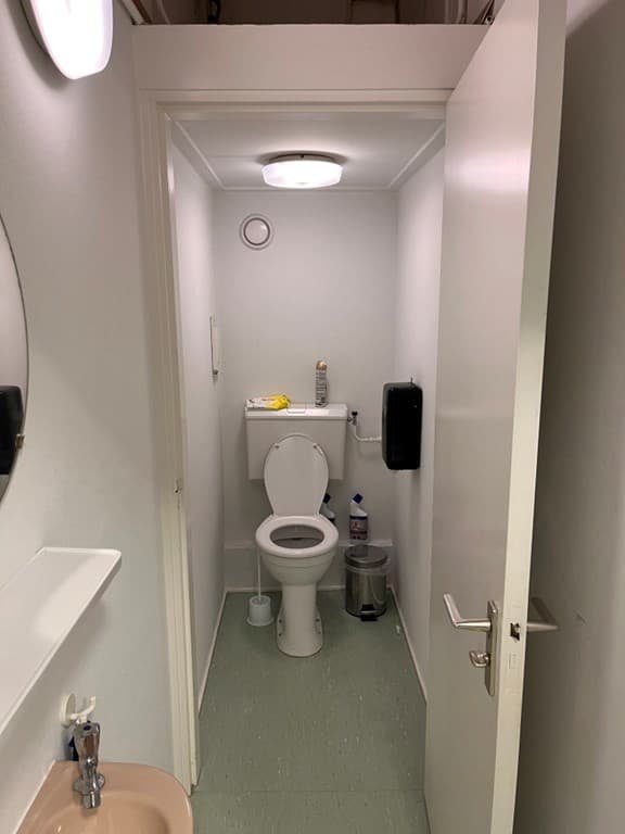 Ingang van een sanitaire ruimte met toilet en wastafel, lichtgrijze muren en groene vloer.