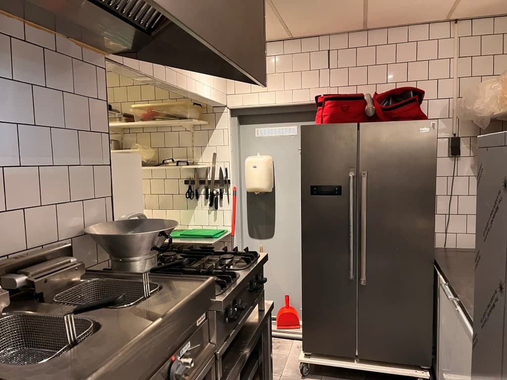 Keuken met RVS apparatuur, gasfornuis, frituurpan, koelkast en witte tegels op de muren.