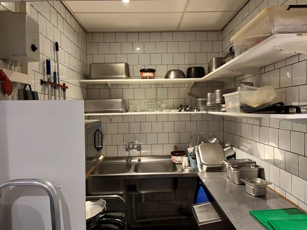 Keuken met witte tegels, metalen rekken met keukengerei en kookpotten, een spoelbak en plastic opbergdozen