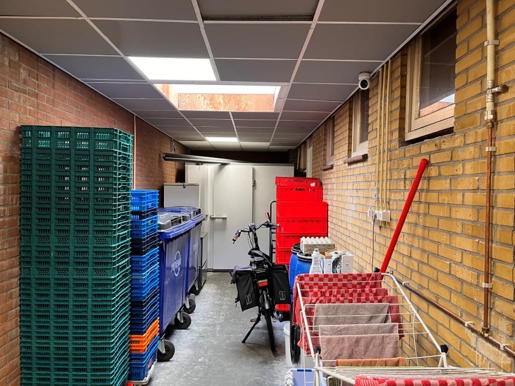 Ruime gang met stapels plastic bakken, fietsen en een wasrek in een gedeeltelijk verlichte ruimte